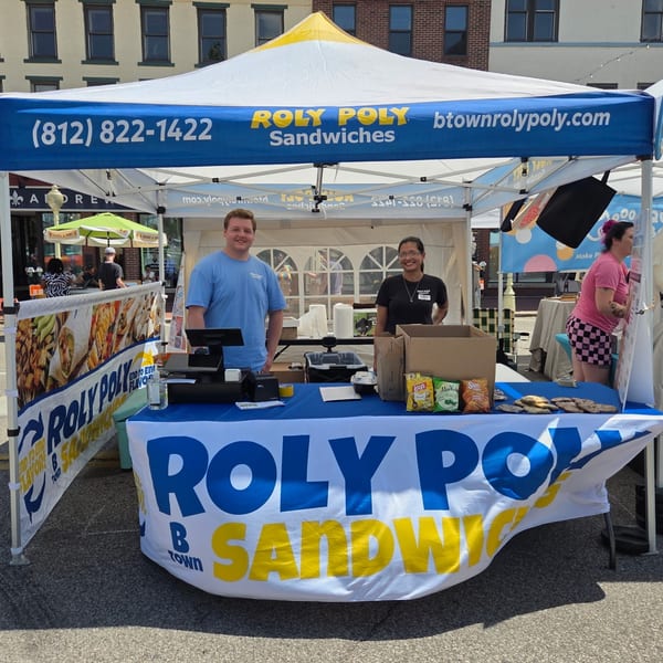 Two Roly Poly Sandwiches employees stand behind a branded tent and table at an outdoor event.