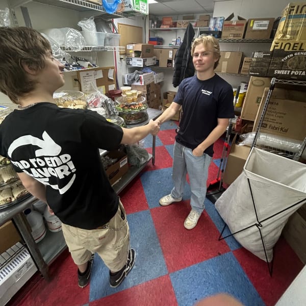 Two young men shaking hands in a Roly Poly sandwich shop kitchen with shelves of supplies.