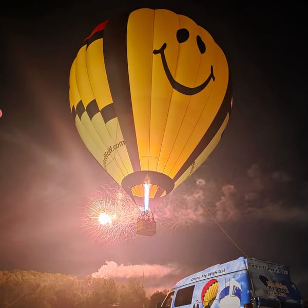 A yellow hot air balloon with a smiley face glows at night, with fireworks and a branded van below.
