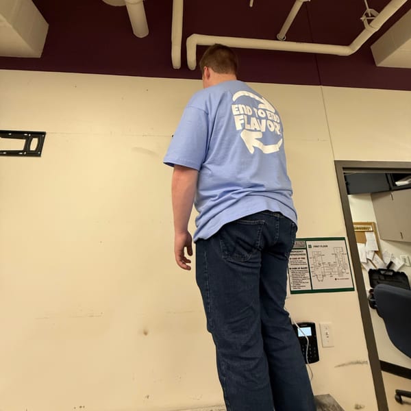 A person wearing a blue "End to End Flavor" t-shirt stands facing a wall in an office setting.