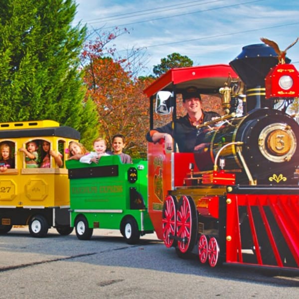 A conductor drives a colorful Roly Poly Express train with children waving from the passenger cars.
