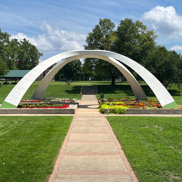 A large white archway sculpture stands over a pathway, surrounded by vibrant flower beds and green grass.
