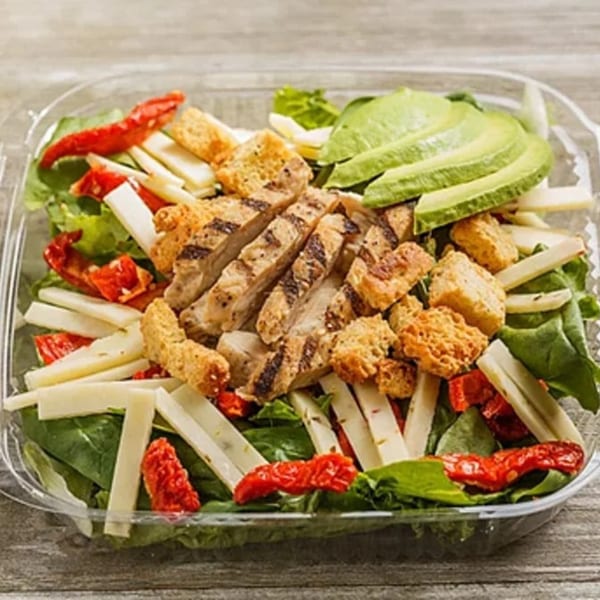 Grilled chicken salad with avocado, croutons, cheese, and sun-dried tomatoes in a plastic container.