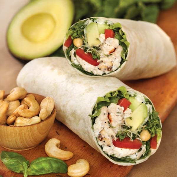 Two Roly Poly chicken salad wraps with avocado, tomato, and lettuce, next to cashews and basil.