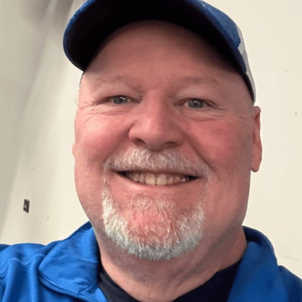 A smiling man with a gray beard and blue cap, wearing a blue jacket.