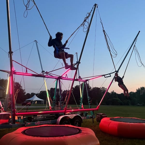 Two people bungee jumping on a trampoline ride with neon lights at dusk.