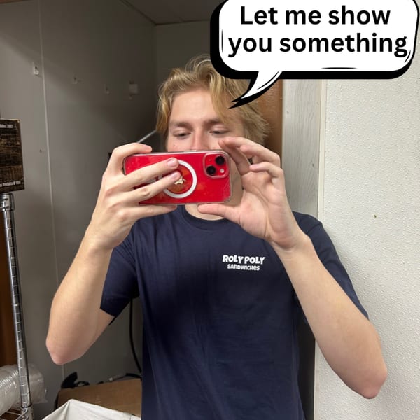 A young man in a Roly Poly Sandwiches shirt holds up a red phone to take a picture.