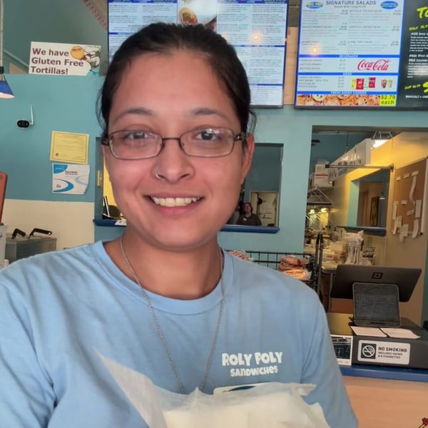 Smiling Roly Poly Sandwiches employee in glasses and light blue shirt stands in front of menu.